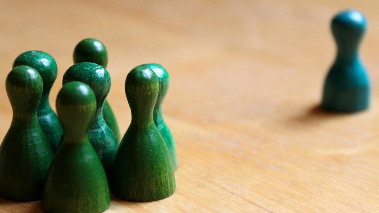 Foto: mehrere Holzspielfiguren, die zusammen als Gruppe stehen gegenüber einer einzelnen alleine stehenden Holzfigur