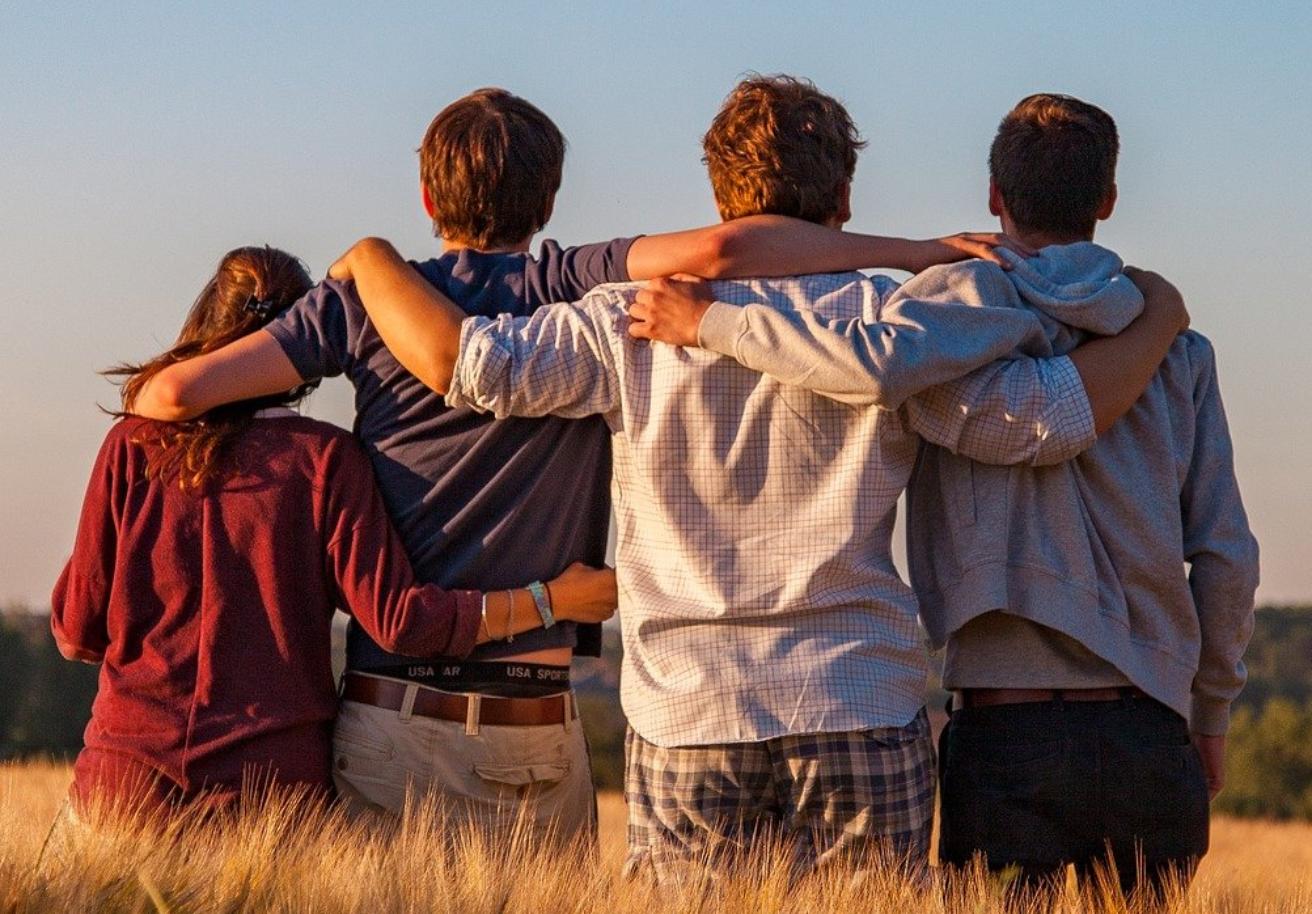 Foto: vier Freunde / Jugendliche umarmen sich, stehen auf einem Feld, von hinten fotografiert