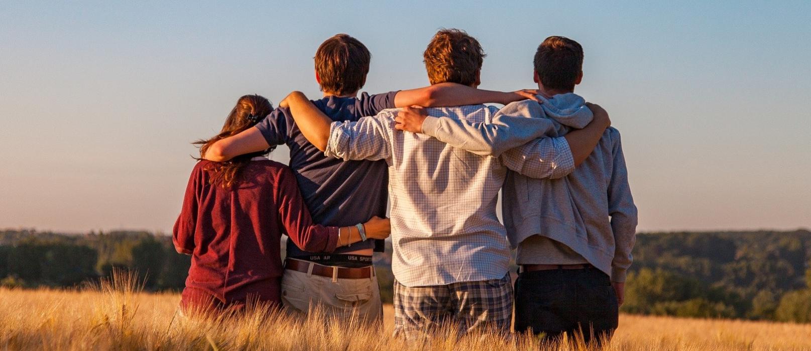 Foto: vier Freunde / Jugendliche umarmen sich, stehen auf einem Feld, von hinten fotografiert