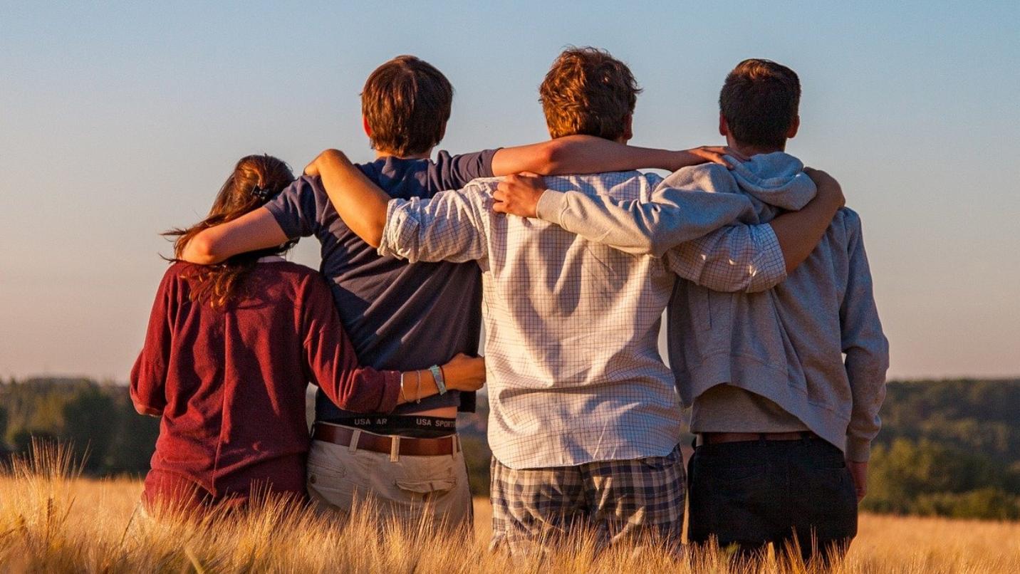 Foto: vier Freunde / Jugendliche umarmen sich, stehen auf einem Feld, von hinten fotografiert
