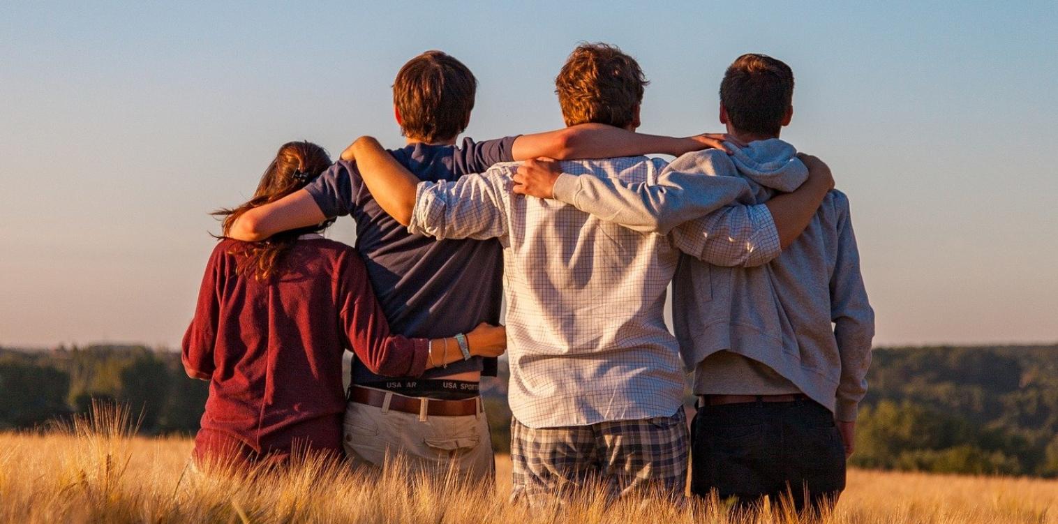 Foto: vier Freunde / Jugendliche umarmen sich, stehen auf einem Feld, von hinten fotografiert